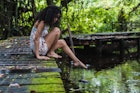 Woman with curly hair sitting on bridge stretches out leg towards water in green forest in Bocas del Toro Islands, Panama.